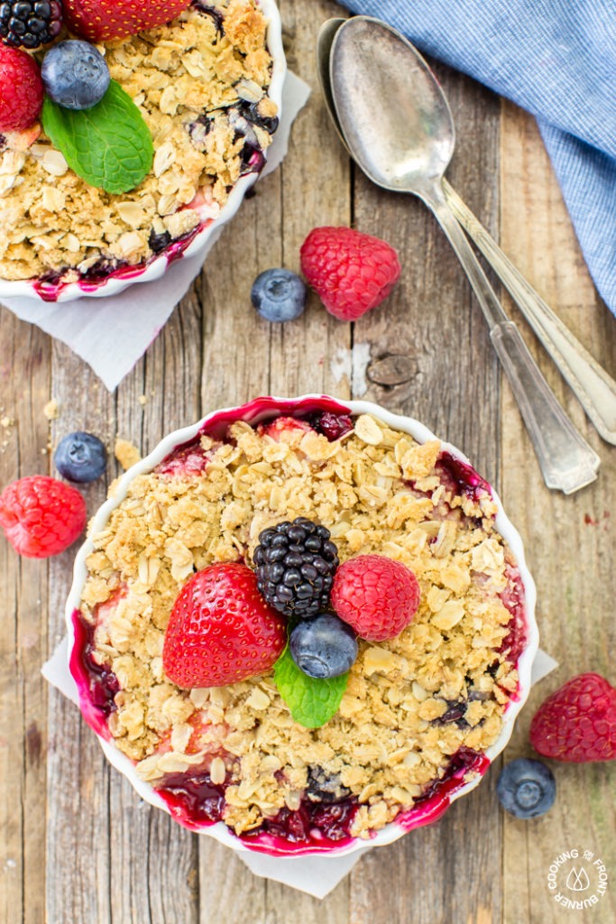 Overhead flat-lay of mixed berry crisp ingredients — fresh strawberries, blueberries, raspberries, rolled oats, brown sugar, cold butter cubes, cinnamon, and cornstarch arranged on a white marble surface