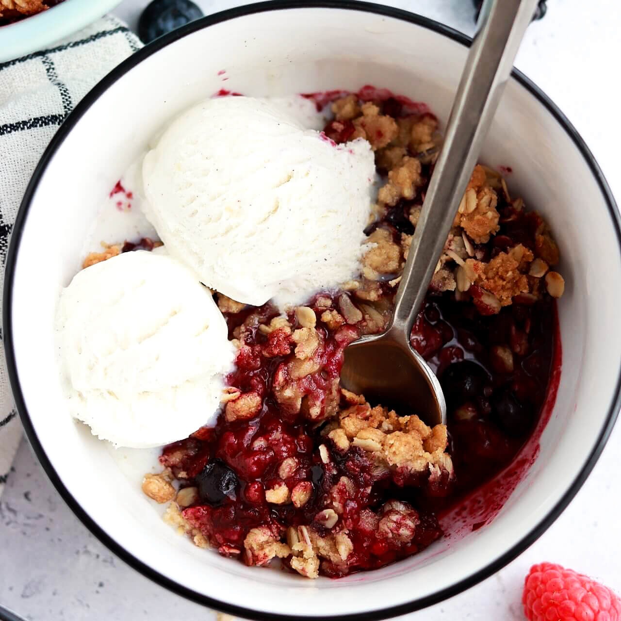 Close-up of a single serving of Mixed Berry Crisp in a white bowl with a scoop of vanilla ice cream, showing the jammy purple-red berry filling and golden oat crumble topping, photographed from above