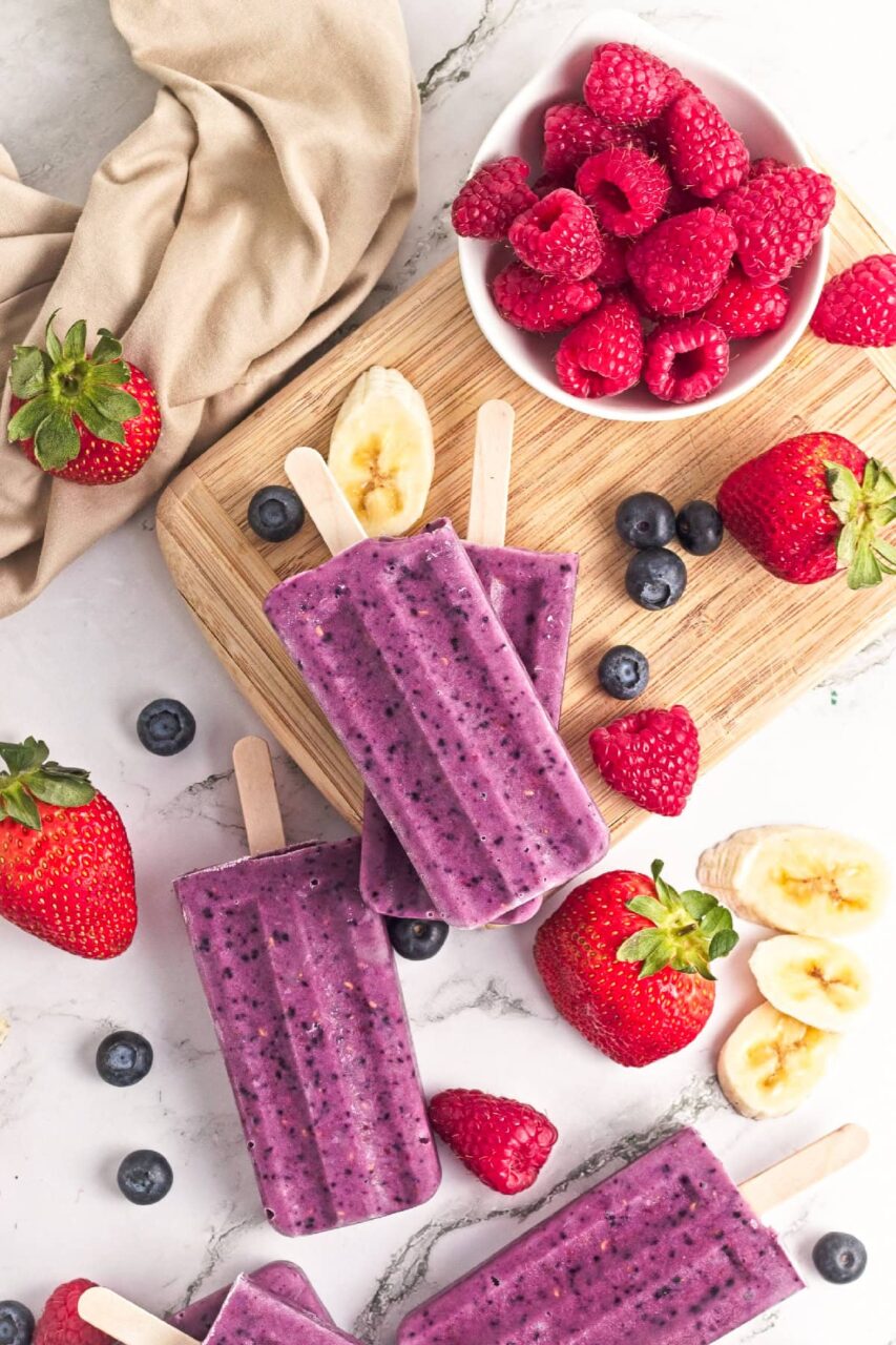 Overhead flat-lay of mixed berry popsicle ingredients including fresh strawberries, blueberries, raspberries, honey, lemon, and coconut milk on a white marble surface