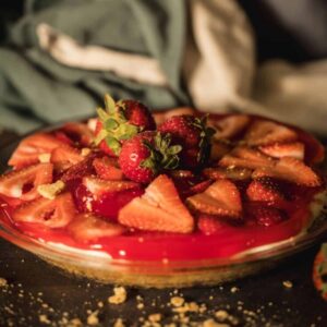 No bake cheesecake on a white cake stand with fresh strawberries and a buttery graham cracker crust, sliced to show the thick creamy filling