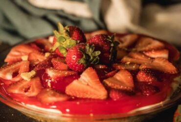 No bake cheesecake on a white cake stand with fresh strawberries and a buttery graham cracker crust, sliced to show the thick creamy filling