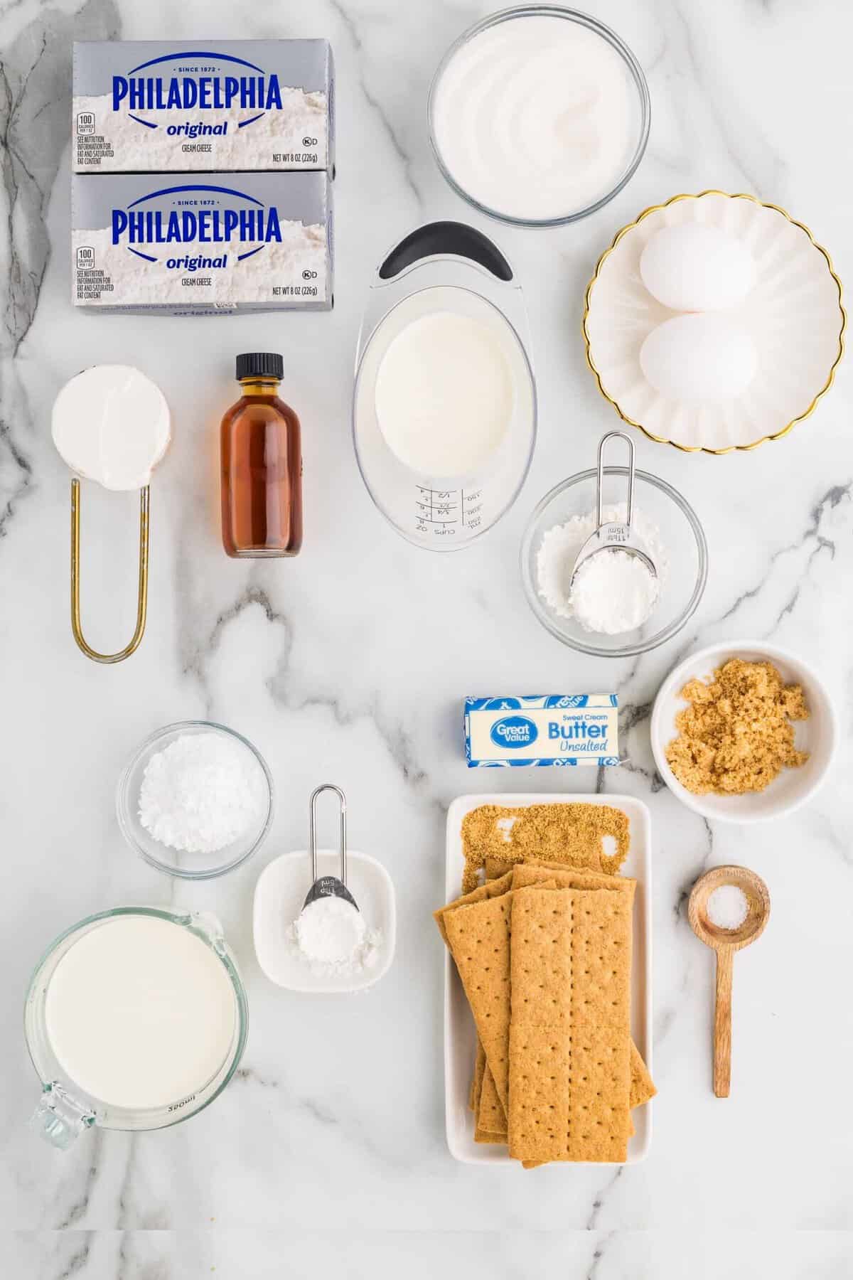 Overhead flat-lay of no bake cheesecake ingredients including cream cheese blocks, graham crackers, heavy cream, powdered sugar, vanilla, lemon, and butter on a white marble surface