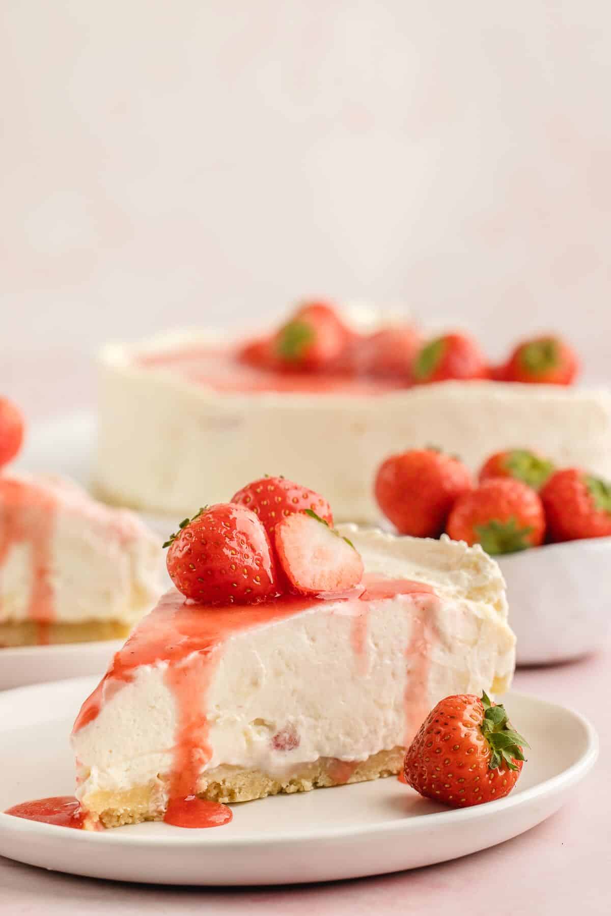 Close-up of a single slice of no bake cheesecake on a white plate topped with fresh strawberry slices and a drizzle of strawberry sauce, showing the dense creamy filling and golden graham cracker crust