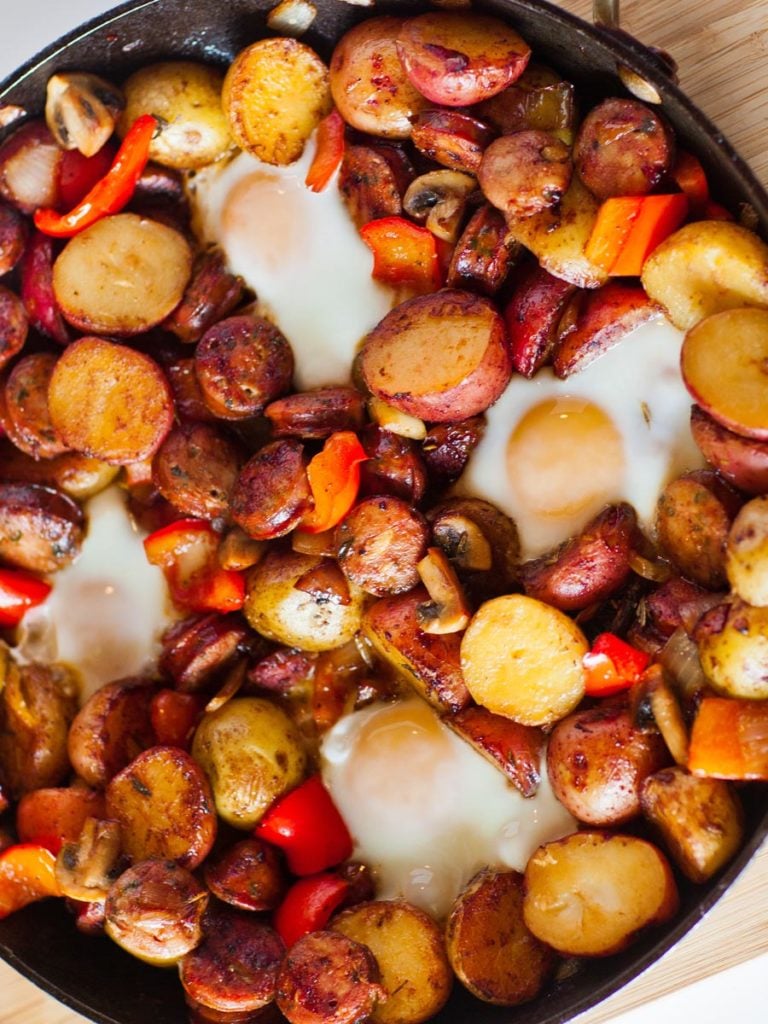 Diced potatoes, sliced peppers, raw eggs, and breakfast sausage laid out as ingredients prep shot beside a cast iron skillet on a rustic wooden surface