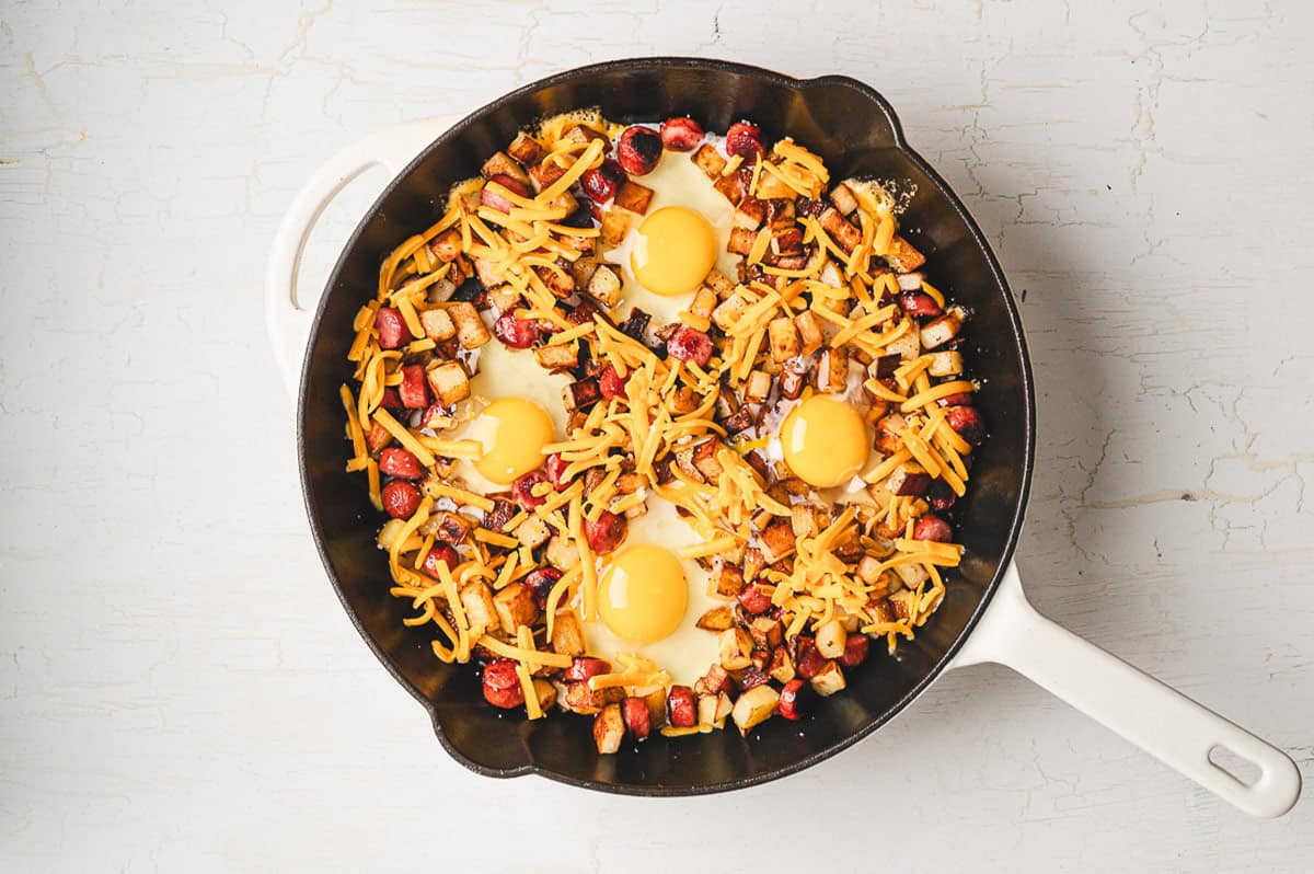 Close-up of one pan camping breakfast skillet with a runny egg yolk breaking over crispy potato hash and sausage, garnished with fresh chives, cast iron skillet visible