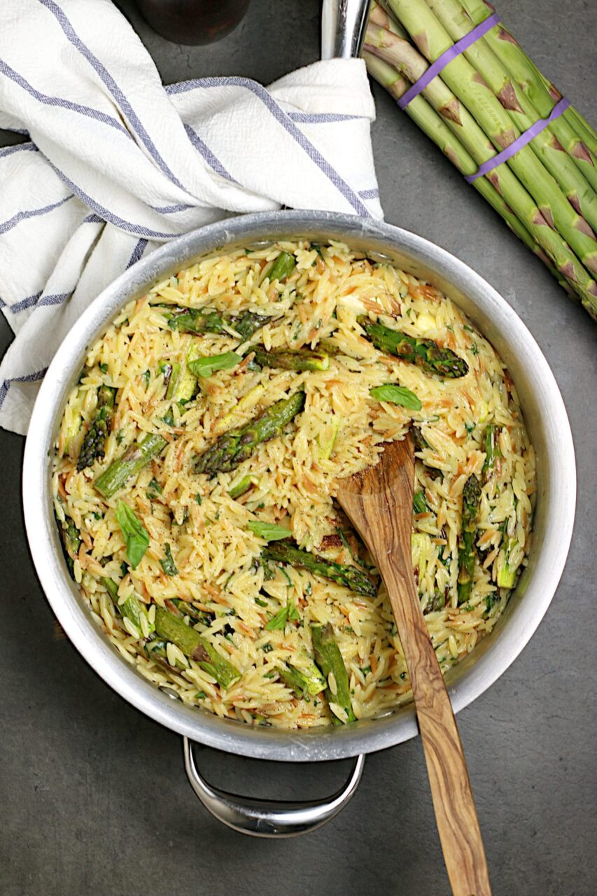 Overhead flat lay of ingredients for one pot cheesy orzo — dry orzo, chicken broth, fresh asparagus spears, shallot, garlic cloves, block of Parmesan, and a stick of butter on a light wooden surface
