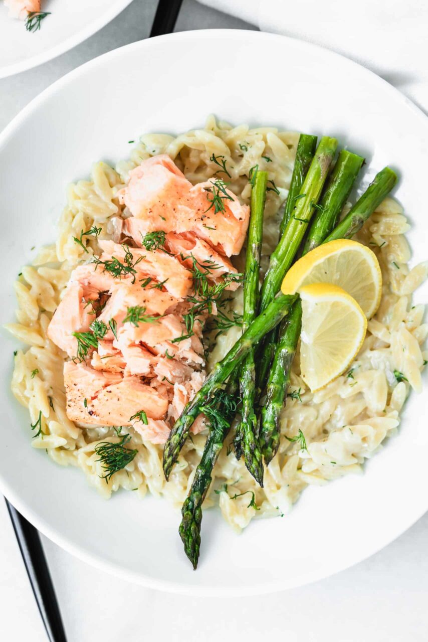 Close-up shot of creamy cheesy orzo with asparagus in a white bowl, steam rising, Parmesan shaved on top, lemon wedge on the side, rustic linen napkin underneath