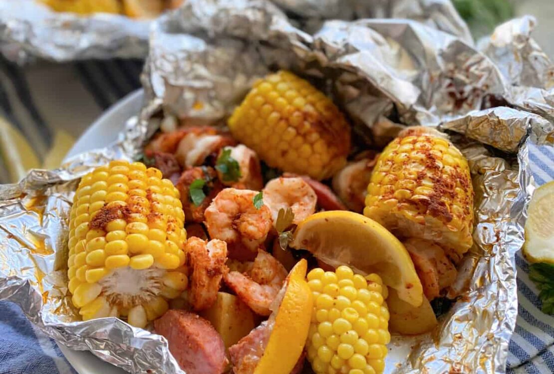 Shrimp boil foil packets opened on a wooden board showing shrimp, smoked sausage, corn, and potatoes with melted butter and parsley