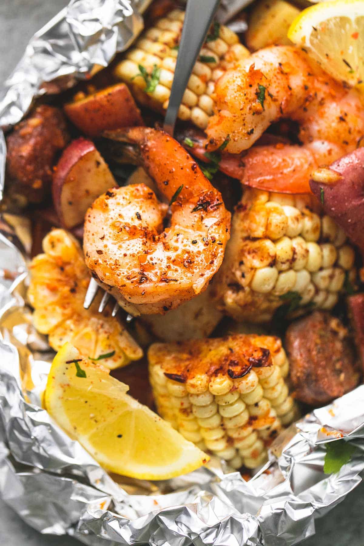 Close-up of opened shrimp boil foil packet showing plump shrimp, sliced sausage, and corn glistening with Cajun butter sauce, garnished with parsley and lemon wedges
