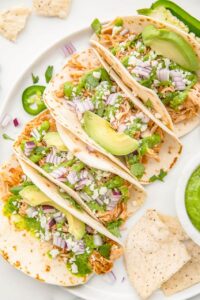Slow cooker chicken tacos served in warm flour tortillas with shredded cabbage, sour cream, fresh cilantro, lime wedges, and diced avocado on a rustic wooden board