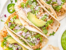 Slow cooker chicken tacos served in warm flour tortillas with shredded cabbage, sour cream, fresh cilantro, lime wedges, and diced avocado on a rustic wooden board