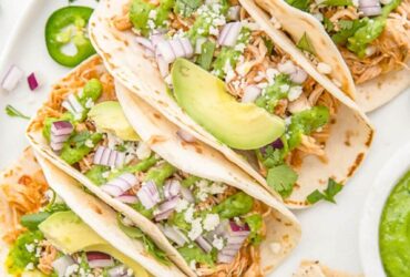 Slow cooker chicken tacos served in warm flour tortillas with shredded cabbage, sour cream, fresh cilantro, lime wedges, and diced avocado on a rustic wooden board