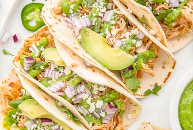 Slow cooker chicken tacos served in warm flour tortillas with shredded cabbage, sour cream, fresh cilantro, lime wedges, and diced avocado on a rustic wooden board