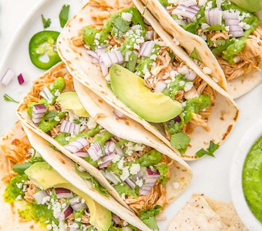 Slow cooker chicken tacos served in warm flour tortillas with shredded cabbage, sour cream, fresh cilantro, lime wedges, and diced avocado on a rustic wooden board