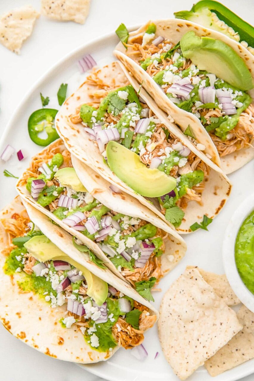 Slow cooker chicken tacos served in warm flour tortillas with shredded cabbage, sour cream, fresh cilantro, lime wedges, and diced avocado on a rustic wooden board