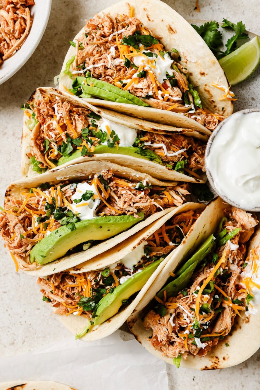 Overhead flat lay of slow cooker chicken taco ingredients — raw chicken thighs, jar of salsa, taco seasoning packet, cumin, smoked paprika, chicken broth, garlic, and lime arranged on a white marble surface