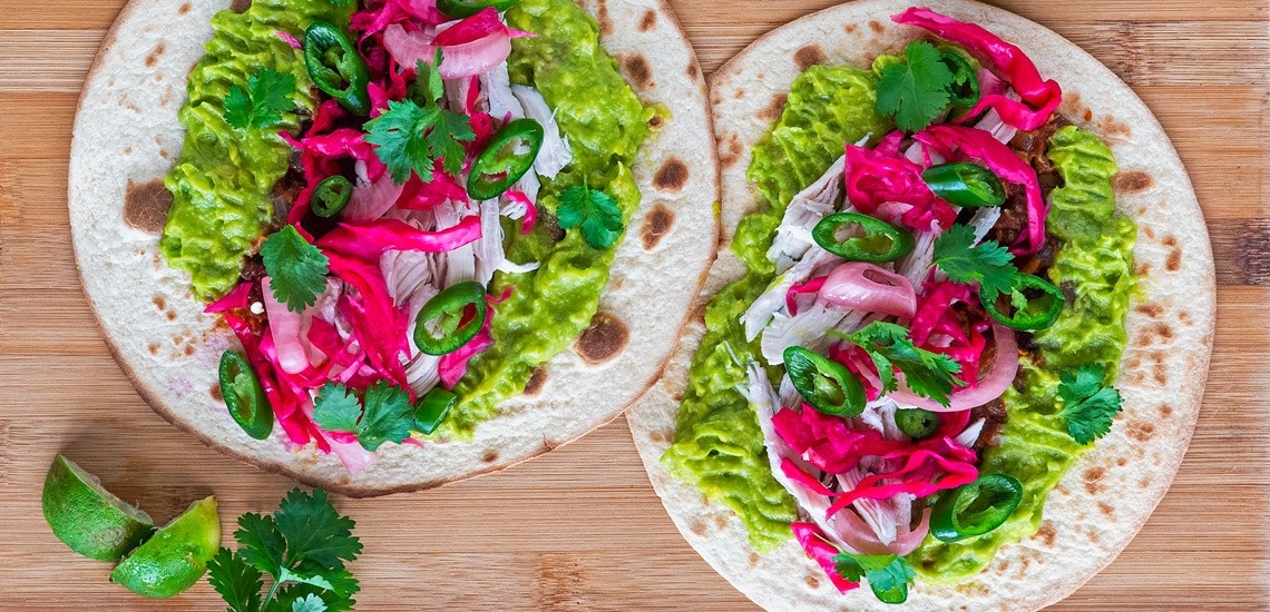 Close-up of two slow cooker chicken tacos in corn tortillas topped with cotija cheese, pickled red onions, fresh cilantro, and a squeeze of lime with a vibrant orange-red chicken filling visible