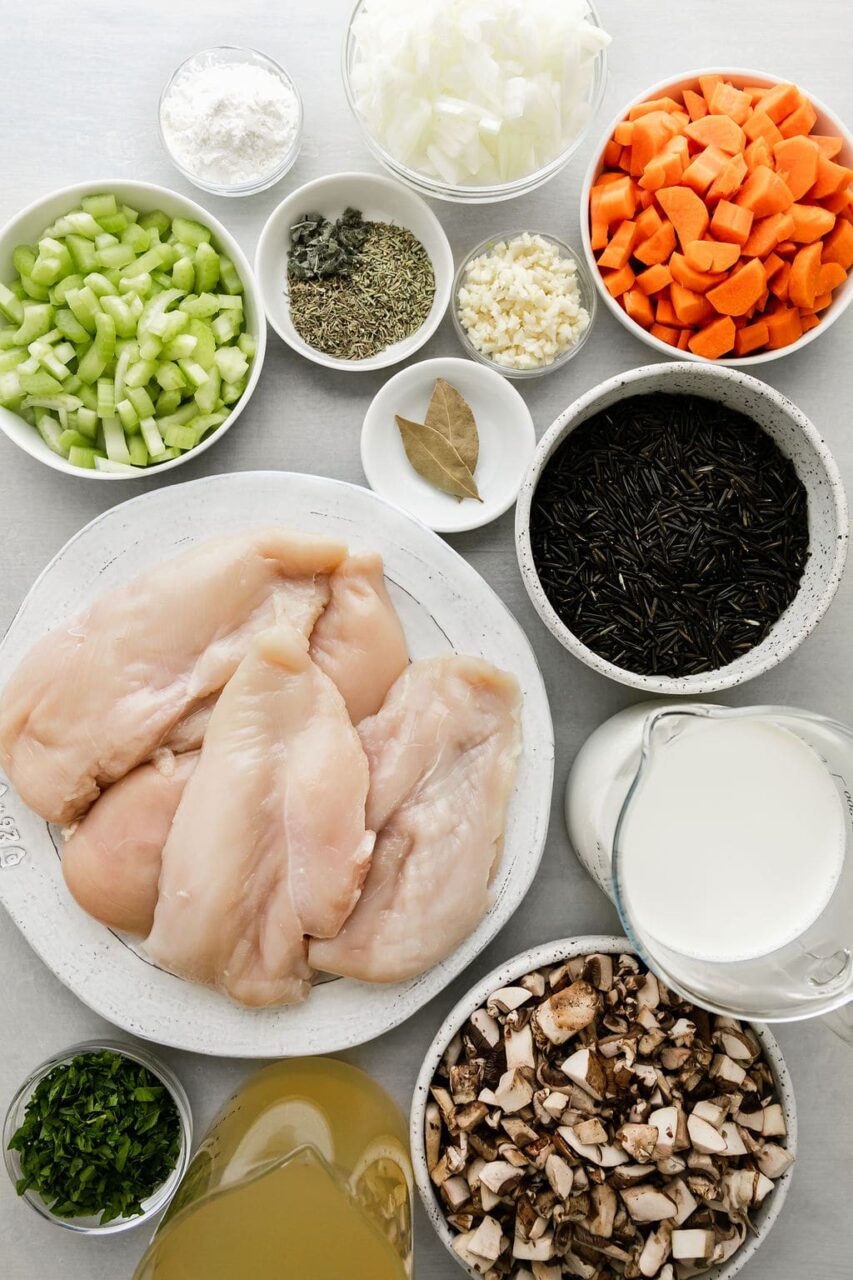 Overhead flat lay of slow cooker chicken wild rice soup ingredients including wild rice, raw chicken thighs, diced carrots, celery, onion, garlic, and broth measured out in bowls on a wooden surface
