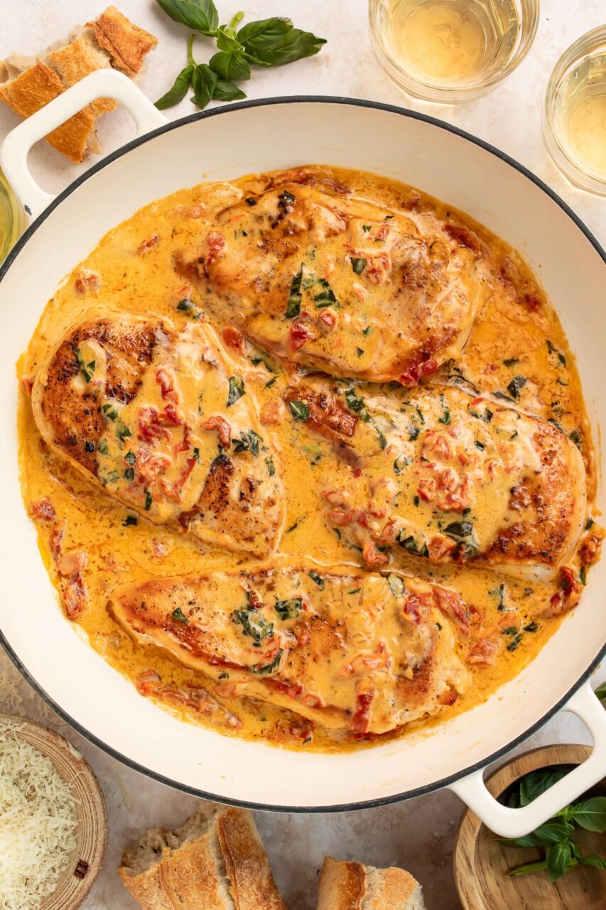 Overhead flat-lay of Slow Cooker Marry Me Chicken ingredients on a wooden cutting board — raw chicken thighs, sun-dried tomatoes in a small bowl, garlic cloves, block of cream cheese, Parmesan, and small bowls of dried spices