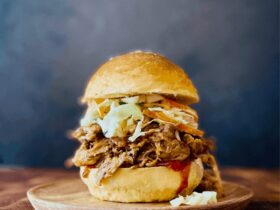 Slow cooker pulled pork piled high on a toasted brioche bun with coleslaw and pickles, served on a wooden board with a side of extra BBQ sauce