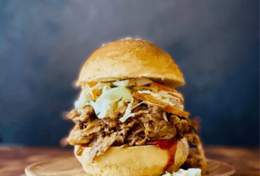 Slow cooker pulled pork piled high on a toasted brioche bun with coleslaw and pickles, served on a wooden board with a side of extra BBQ sauce