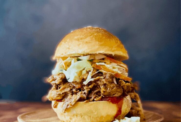 Slow cooker pulled pork piled high on a toasted brioche bun with coleslaw and pickles, served on a wooden board with a side of extra BBQ sauce
