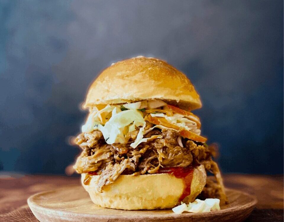 Slow cooker pulled pork piled high on a toasted brioche bun with coleslaw and pickles, served on a wooden board with a side of extra BBQ sauce