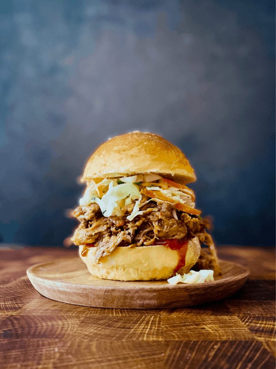 Slow cooker pulled pork piled high on a toasted brioche bun with coleslaw and pickles, served on a wooden board with a side of extra BBQ sauce