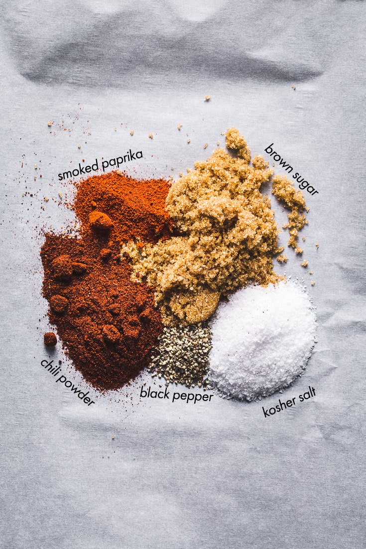 Overhead shot of raw pork shoulder coated in dry spice rub on a cutting board, surrounded by bowls of smoked paprika, brown sugar, garlic powder, apple cider vinegar, and BBQ sauce