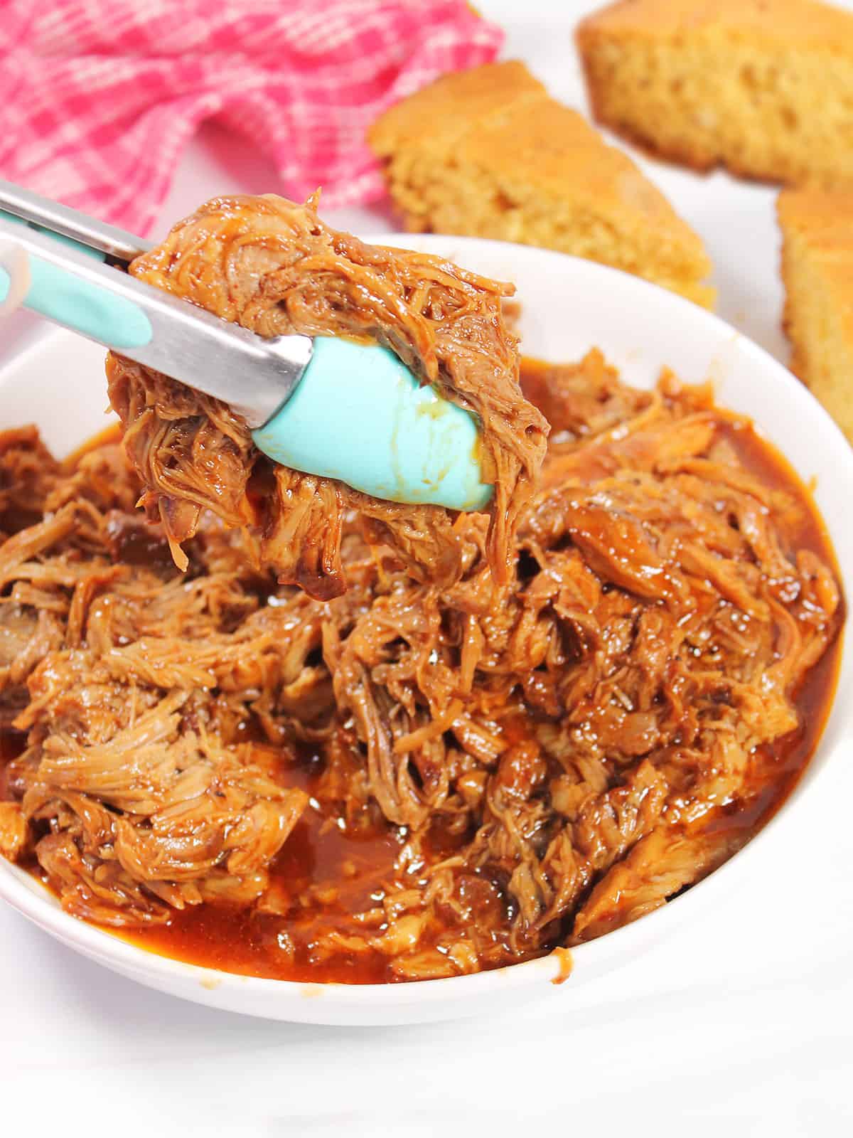 Close-up of pulled pork being lifted with tongs, showing the tender shredded strands glistening with BBQ sauce, with toasted buns and coleslaw blurred in the background