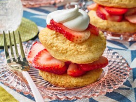 Classic strawberry shortcake with golden biscuits, fresh sliced strawberries, and whipped cream stacked on a white plate, styled on a rustic wood table with a summer backdrop