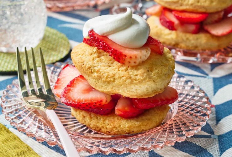 Classic strawberry shortcake with golden biscuits, fresh sliced strawberries, and whipped cream stacked on a white plate, styled on a rustic wood table with a summer backdrop