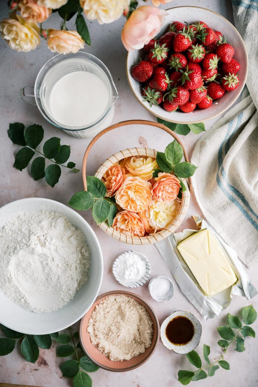 Step-by-step ingredients flat lay for strawberry shortcake — bowl of fresh strawberries, cold butter cubes, flour, sugar, baking powder, and heavy cream arranged on a marble surface
