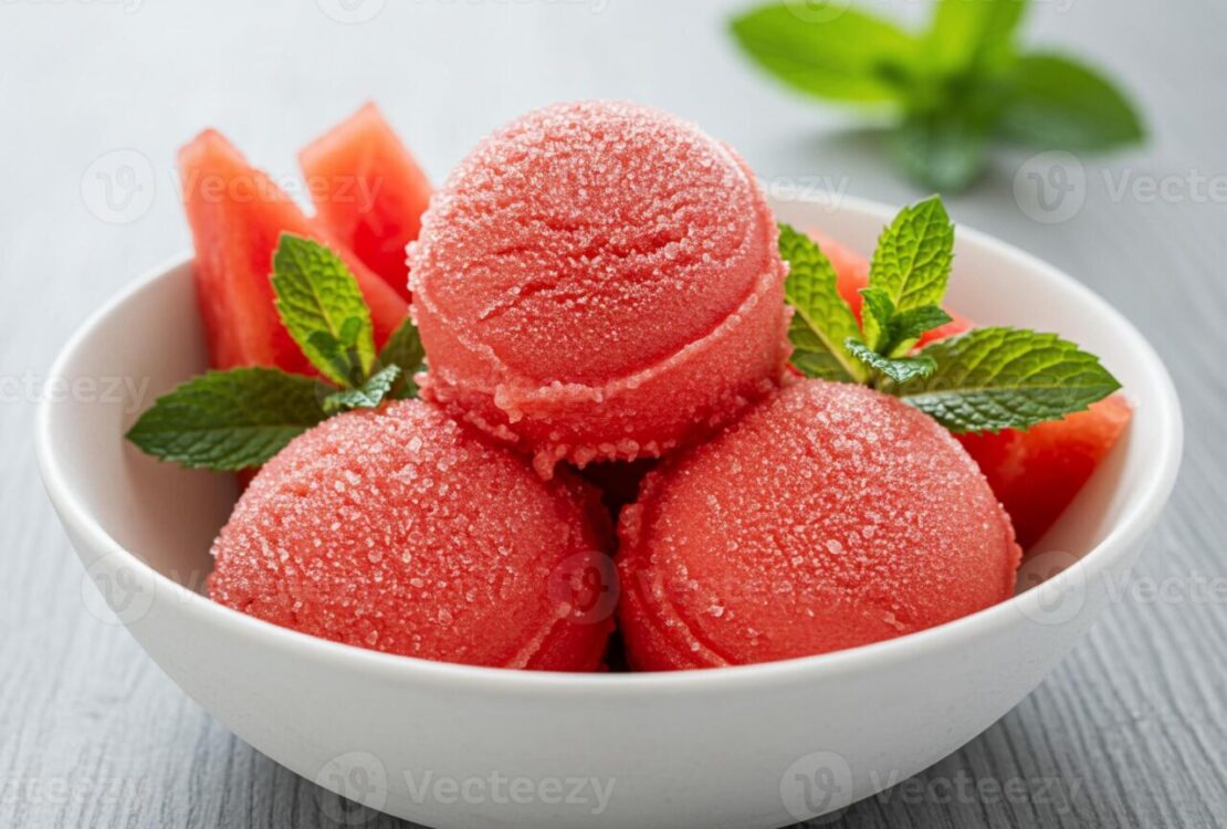 Scoops of vibrant pink watermelon sorbet in a chilled white bowl garnished with fresh mint and a slice of watermelon on a bright summer table