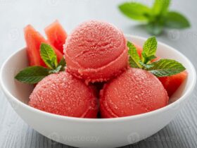 Scoops of vibrant pink watermelon sorbet in a chilled white bowl garnished with fresh mint and a slice of watermelon on a bright summer table