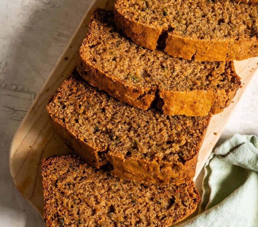 Sliced zucchini bread on a wooden cutting board with a golden-brown crust and moist crumb, warm cinnamon tones, overhead shot