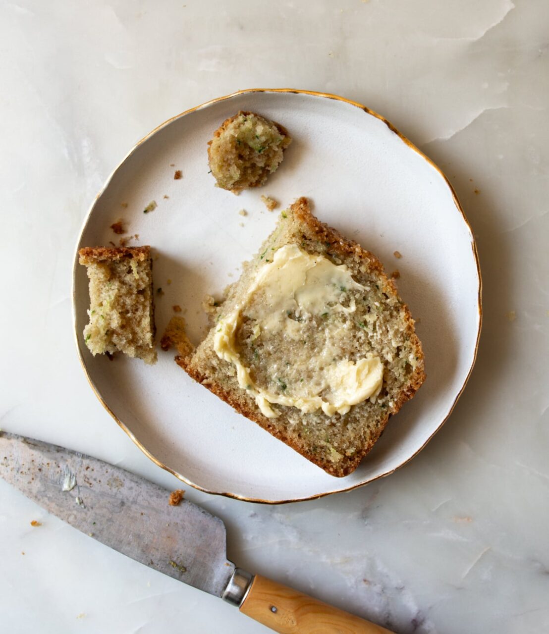Close-up of a thick slice of zucchini bread on a white plate with a pat of butter melting on top, warm natural light, rustic styling