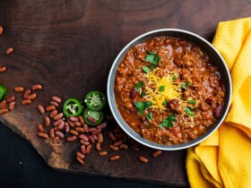 A rustic bowl of thick beef chili topped with shredded cheddar cheese, sour cream, sliced jalapeños, and green onions, photographed overhead on a wooden table
