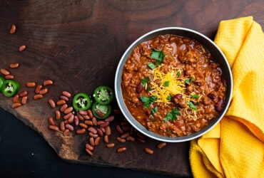 A rustic bowl of thick beef chili topped with shredded cheddar cheese, sour cream, sliced jalapeños, and green onions, photographed overhead on a wooden table