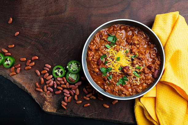 A rustic bowl of thick beef chili topped with shredded cheddar cheese, sour cream, sliced jalapeños, and green onions, photographed overhead on a wooden table
