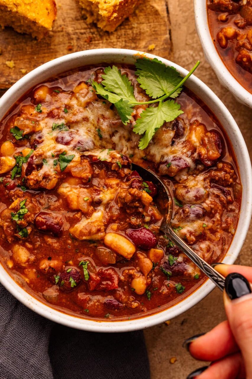 A flat lay of beef chili ingredients including ground beef, canned tomatoes, kidney beans, black beans, spices, onion, garlic, and beef broth arranged on a marble surface