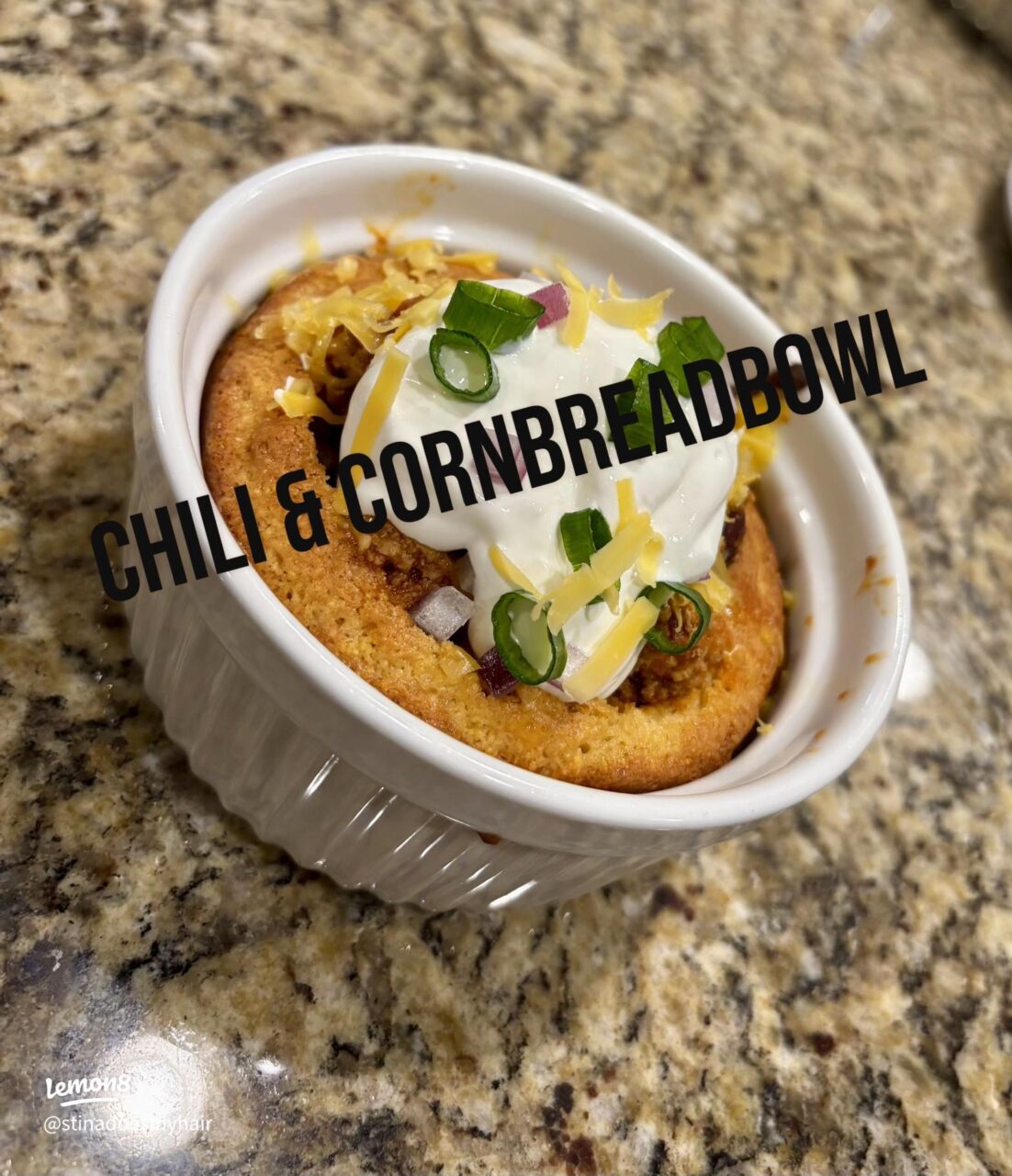 A close-up of a bowl of beef chili served with a side of cornbread, topped with melted cheddar cheese and a dollop of sour cream, shot in warm natural light