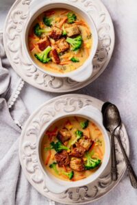 Overhead shot of a white bowl filled with thick, creamy broccoli cheddar soup garnished with shredded cheddar and fresh broccoli florets, served with a crusty bread roll on a rustic wooden table