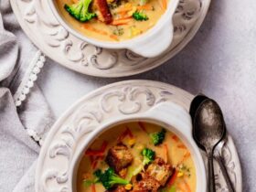 Overhead shot of a white bowl filled with thick, creamy broccoli cheddar soup garnished with shredded cheddar and fresh broccoli florets, served with a crusty bread roll on a rustic wooden table