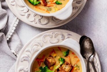 Overhead shot of a white bowl filled with thick, creamy broccoli cheddar soup garnished with shredded cheddar and fresh broccoli florets, served with a crusty bread roll on a rustic wooden table