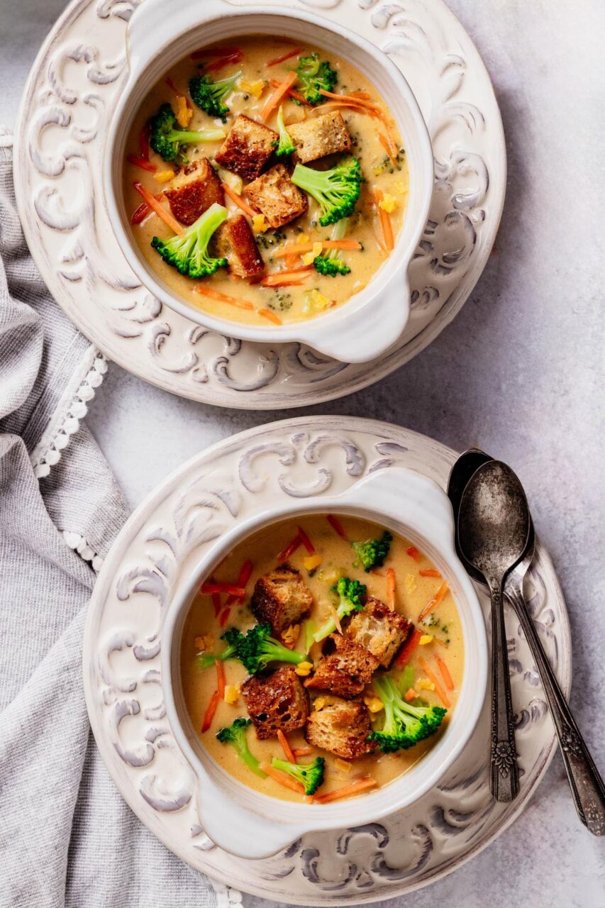 Overhead shot of a white bowl filled with thick, creamy broccoli cheddar soup garnished with shredded cheddar and fresh broccoli florets, served with a crusty bread roll on a rustic wooden table