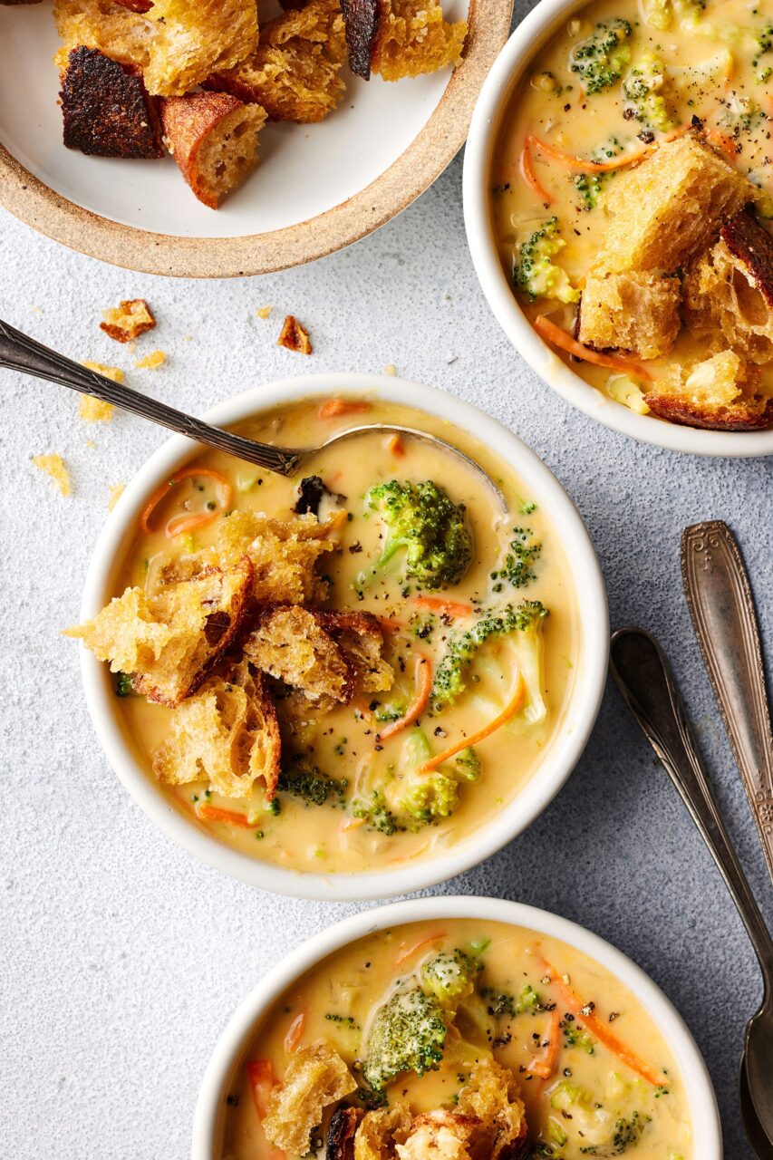 Close-up flat-lay of key ingredients for broccoli cheddar soup including a block of sharp cheddar, fresh broccoli florets, butter, an onion, garlic cloves, flour, and a measuring cup of chicken broth on a light marble surface