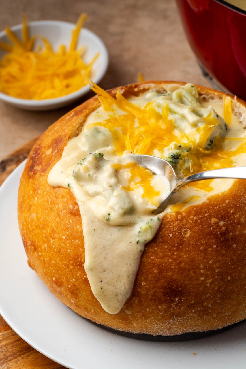 A ladle lifting a scoop of thick, golden broccoli cheddar soup from a Dutch oven, with steam rising and chunks of broccoli and melted cheese visible, with a rustic bread bowl on the side