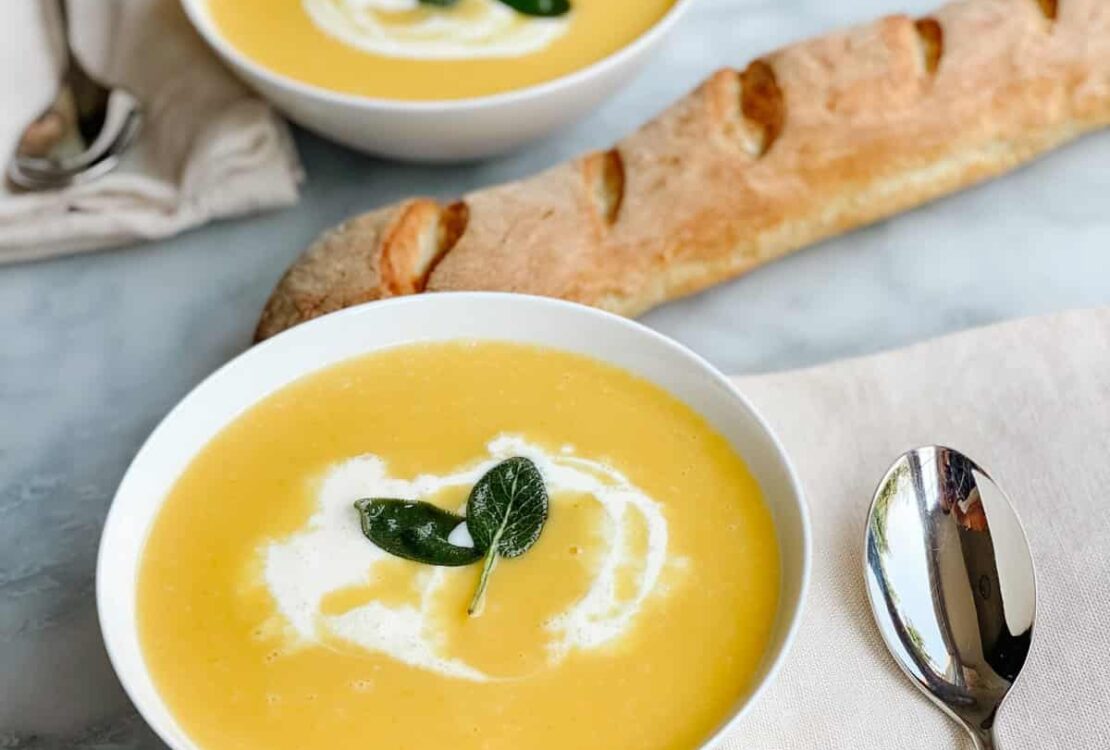 A white bowl of silky butternut squash soup swirled with cream and garnished with crispy sage leaves and a drizzle of olive oil on a wooden table