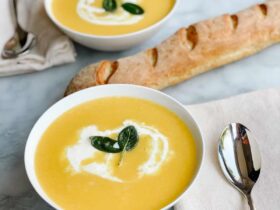 A white bowl of silky butternut squash soup swirled with cream and garnished with crispy sage leaves and a drizzle of olive oil on a wooden table